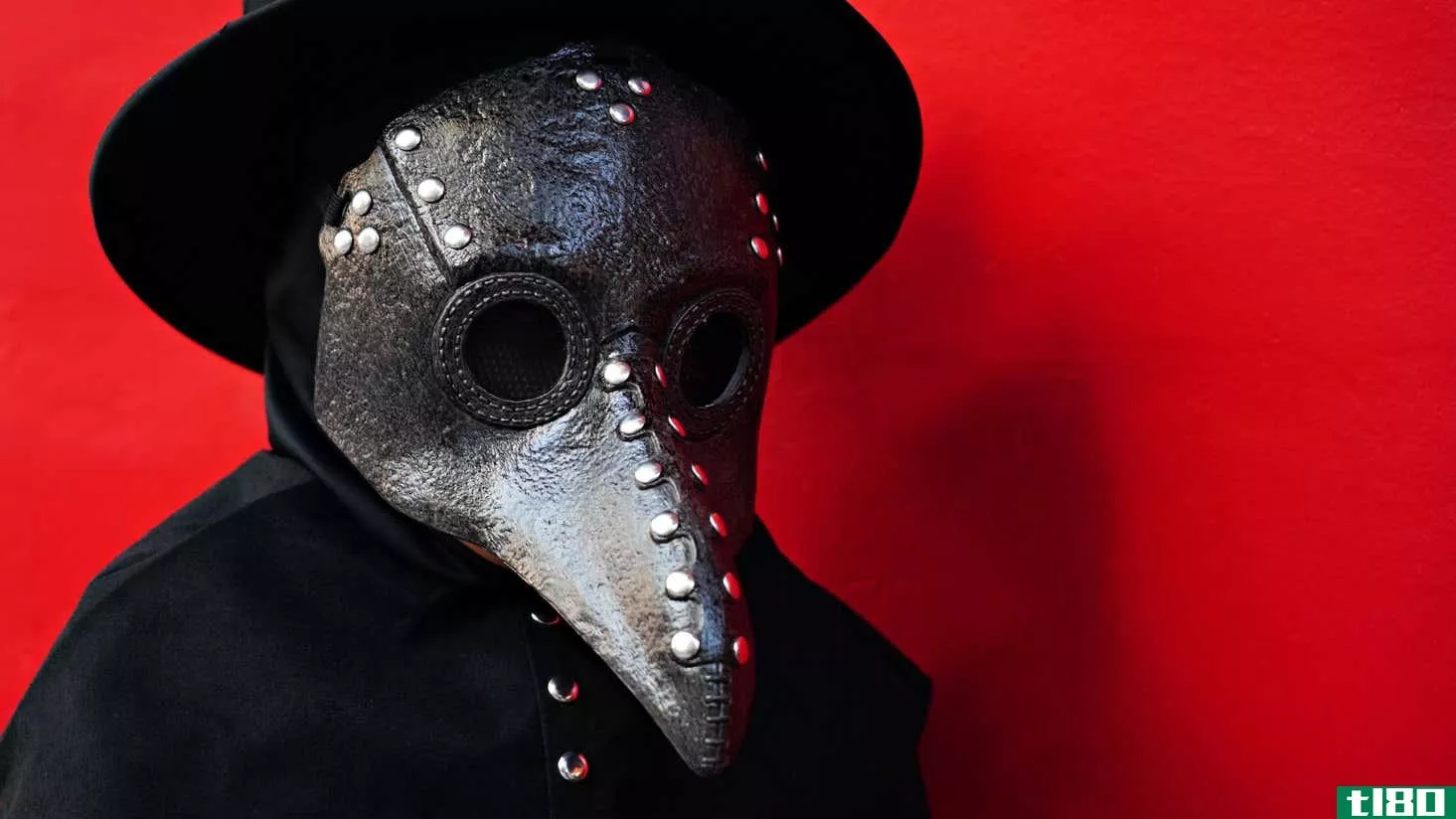 A person dressed in a plague mask and black hat, standing against a red background