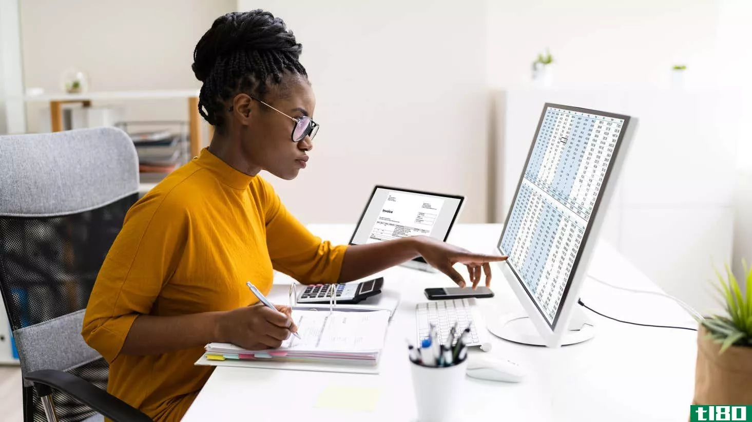 A Black woman sitting at a white desk studies a computer spreadsheet filled with numbers 