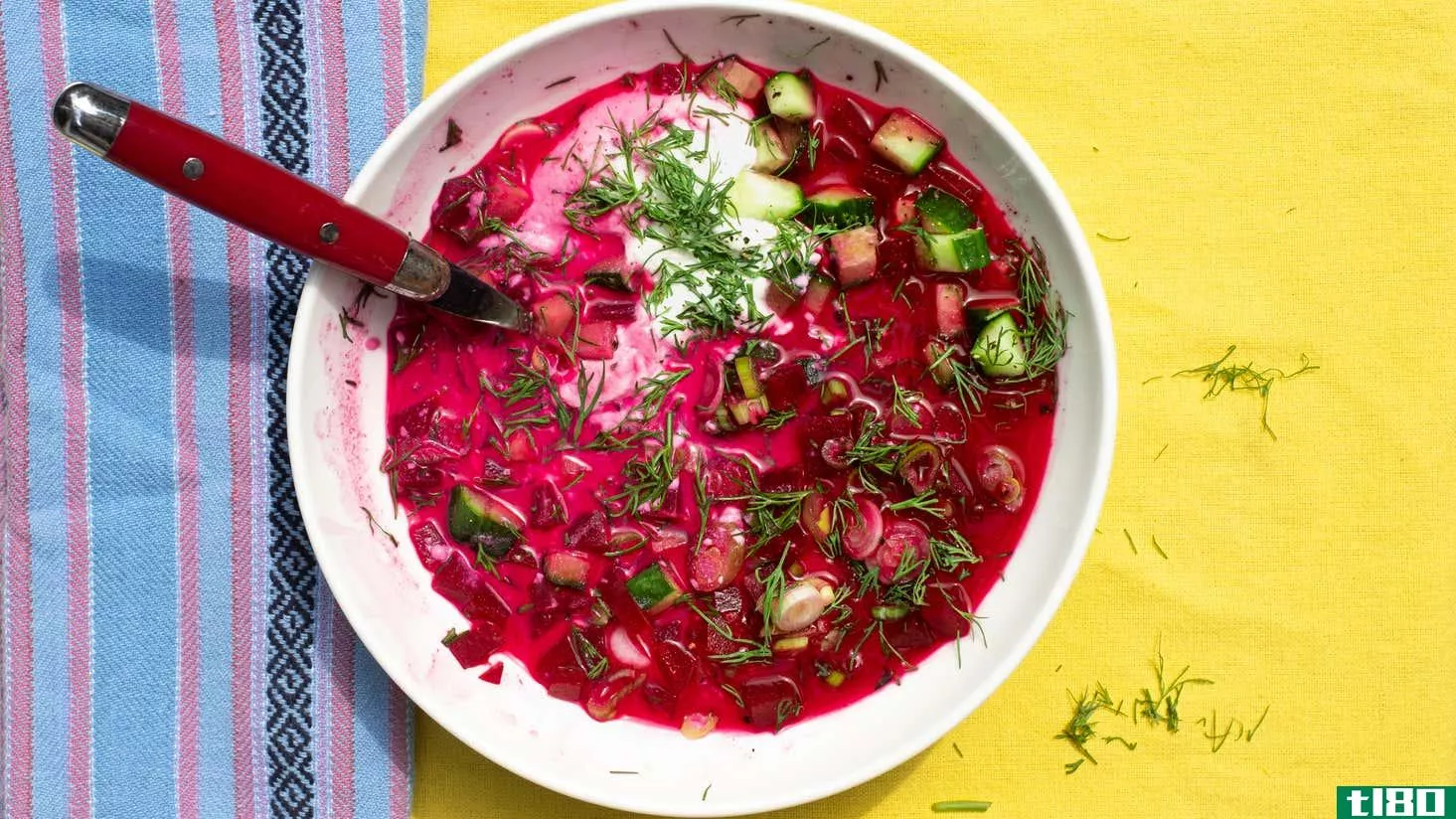 Top-down photograph of a bowl of cold borsch with cucumbers, scallions, dill, and cashew cream. It's in a white bowl on a yellow tablecloth, with a red-handled spoon sticking out of the soup. There's a light blue and pink striped napkin on the left side. 