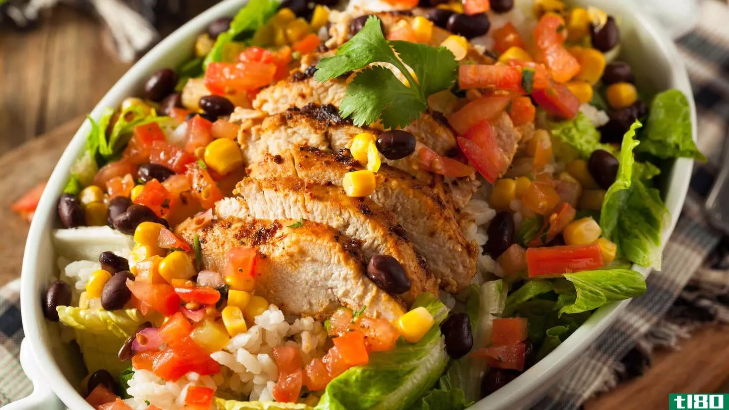 Close-up photo of a Chipotle-style burrito bowl. It has sliced grilled chicken, black beans, corn, rice, lettuce, and tomatoes, with a single cilantro leaf on top for garnish. It's in a white oval bowl on a gray and brown plaid napkin, set on a wooden table.