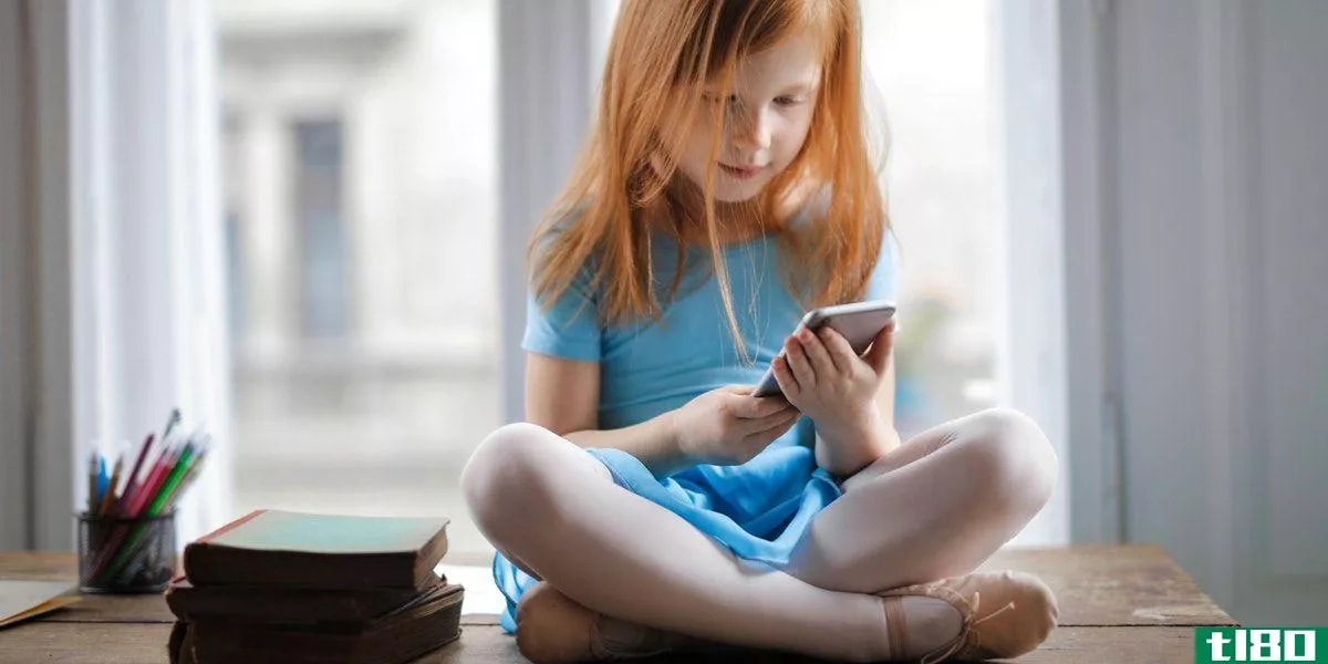 Crossed-legged red-headed girl in a blue dress, holding a phone next to a stack of books and pens