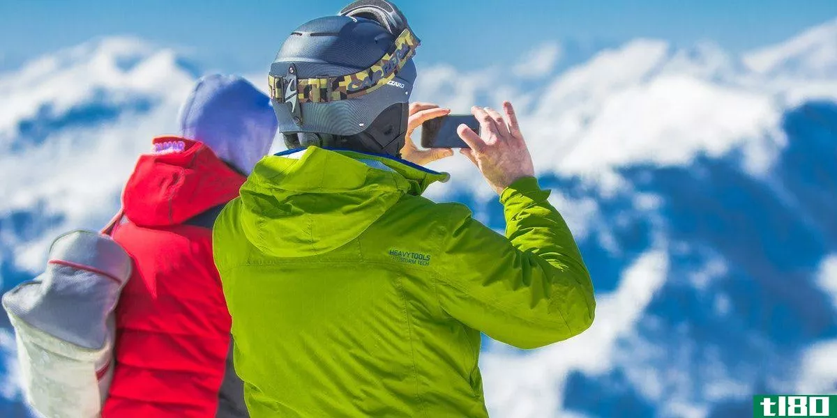 Two people wearing a red and a green jacket with a phone, standing on moutaintop taking a picture