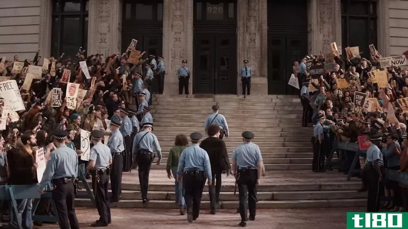 Police officers walk into a courthouse surrounded by protestors in a scene from The Trial of the Chicago 7 