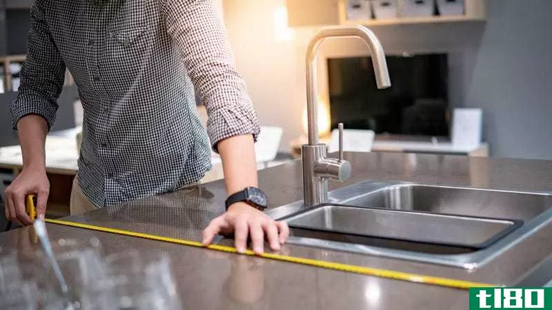 A person wearing a square watch and a navy blue checked shirt uses a yellow tape measure on a dark granite countertop. 