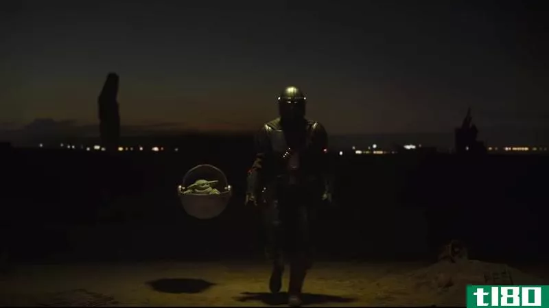 Screenshot from The Mandalorian featuring the titular armord warrior standing near Baby Yoda floating in a pod and surrounded by menacing figures.