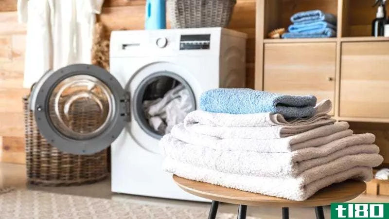 A pile of folded towels sits on a stool in front of an open front-loading washing machine.