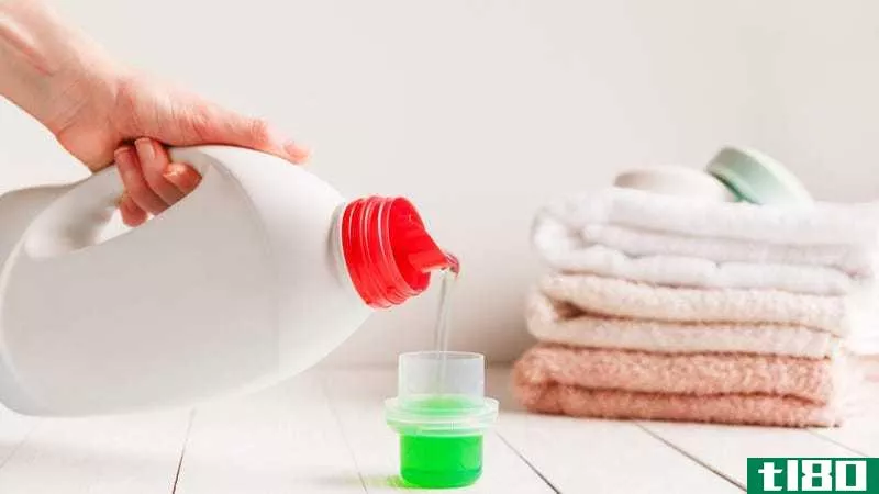 Close-up shot of a person pouring green laundry detergent into a clear cap. There's a pile of folded pink and white towels in the background. 