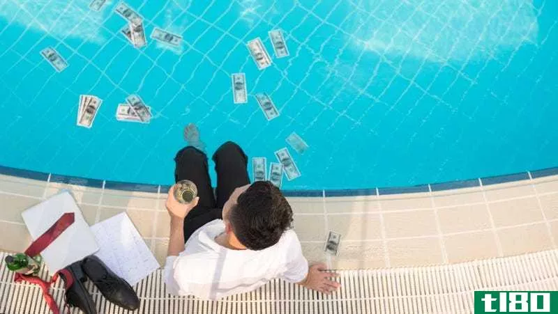 A man sits on the edge of a pool with his feet in the water and a glass in his left hand. There are dollar bills floating in the water.